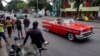 Turistas pasean en un almendrón mientras varias personas esperan por el transporte público en una parada en La Habana.